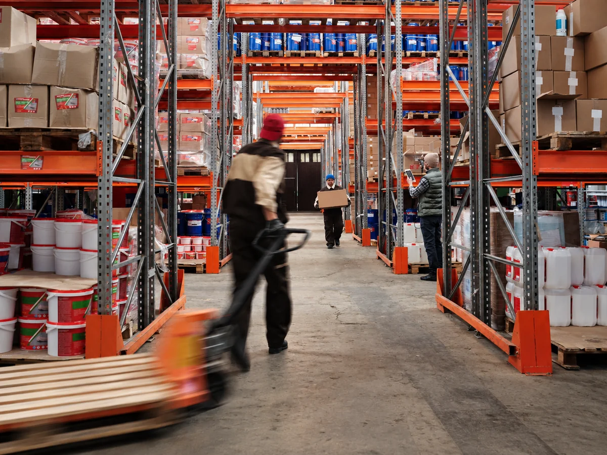 Edited editorial scene of a worker in a red beanie pulling a pallet jack through orange warehouse racking.