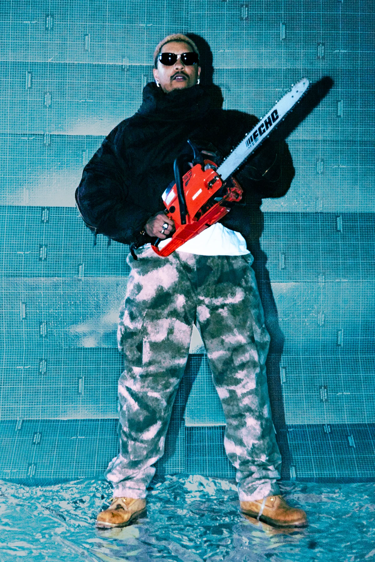 Fashion editorial portrait with camouflage trousers, a dark top and a red chainsaw against turquoise tile.