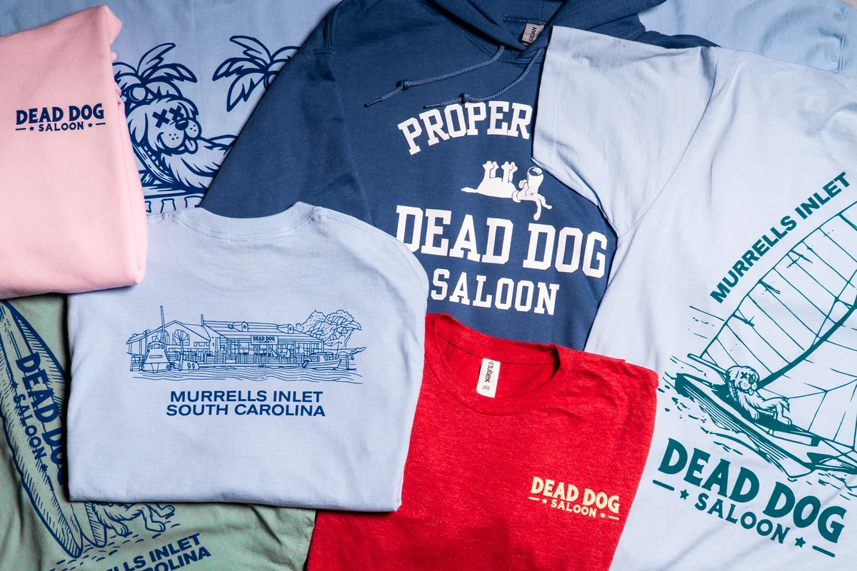 Dead Dog Saloon merchandise arranged with an indigo hoodie, a red shirt and light blue shirts.