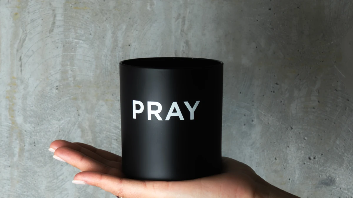 Black PRAY candle supported by a hand against a gray wall.