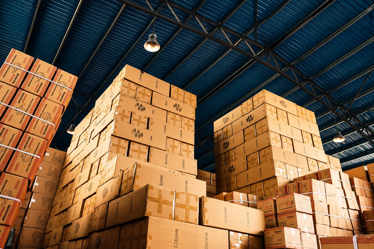 Edited editorial image of stacked cartons beneath a blue warehouse roof.